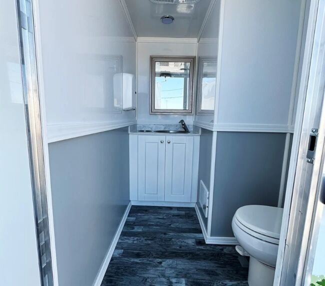 Interior view of 17-foot ADA restroom trailer featuring toilet, vanity sink cabinet, mirror, air vent, and wood-style flooring.