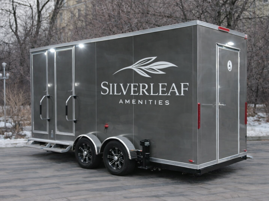 Grey luxury restroom trailer with dual axle wheels and exterior lights parked in an urban outdoor setting during winter.