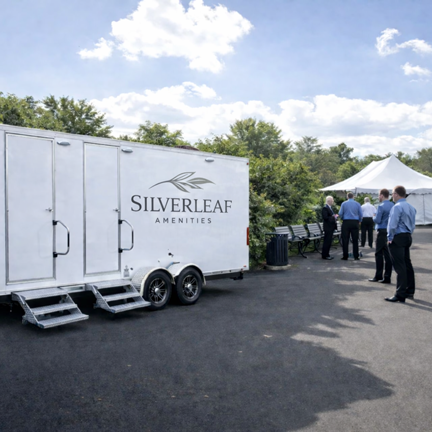 White luxury restroom trailer with dual entry doors and steps set up at an outdoor corporate event with guests gathered nearby.