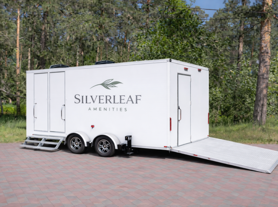 Silverleaf Amenities white restroom trailer with dual side doors, alloy wheels, and an ADA access ramp, parked on a red and gray brick pavement surrounded by tall pine trees and green forest background.