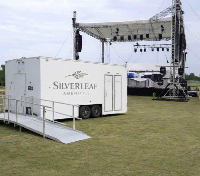 ADA-compliant restroom trailer with ramp at outdoor event near stage setup, available for festival and event rentals in Colorado